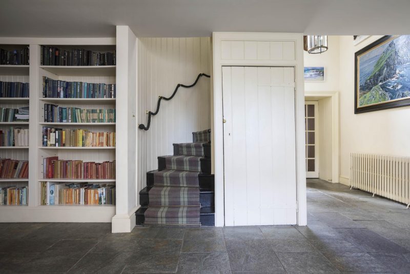 A concealed spiral staircase with tartan carpet, ascends from a flagstoned hall lined with bookshelves and simple, painted panelling. Renovation in a Scottish country house, Isle of Mull.