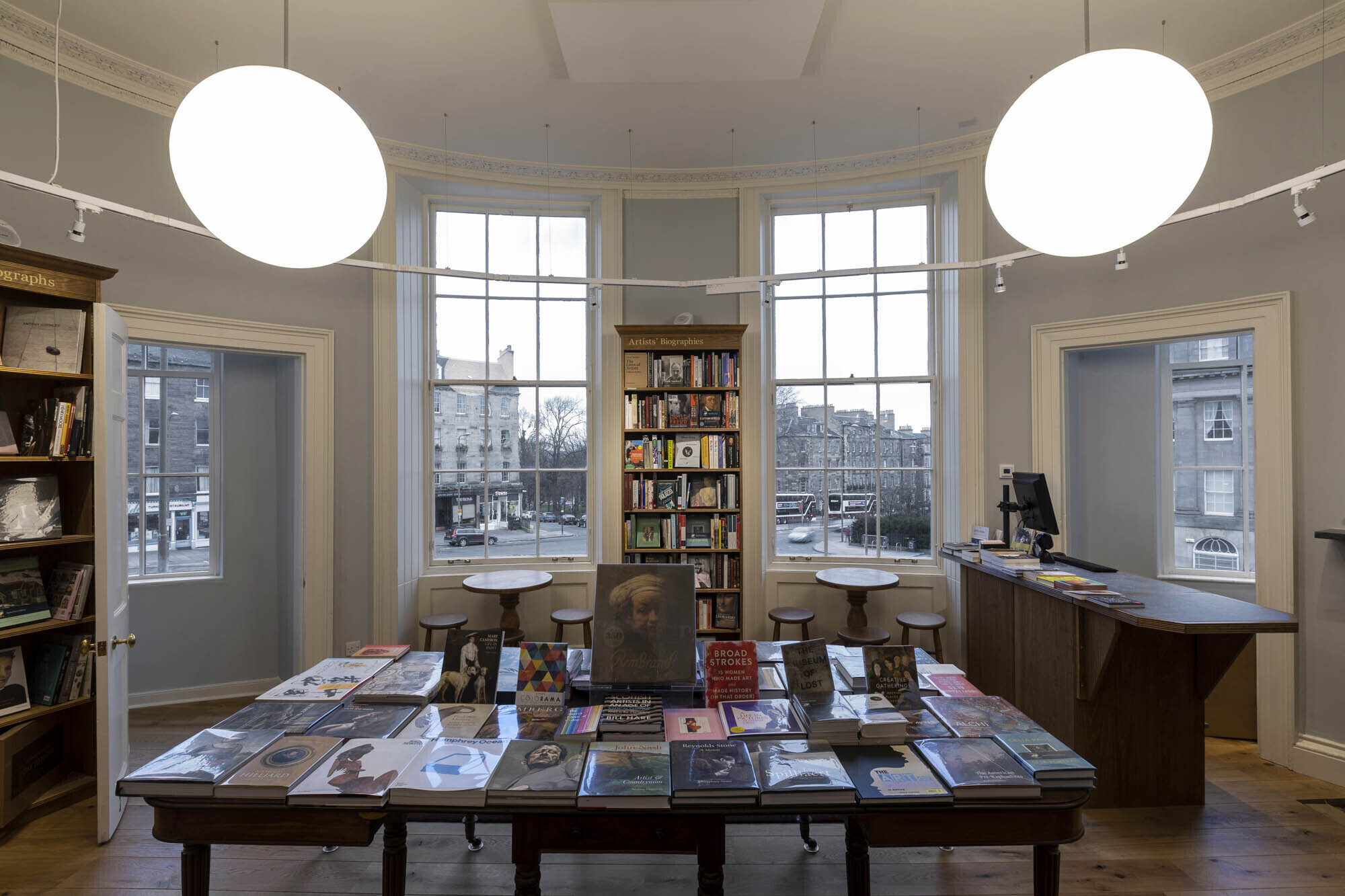 Inside the first floor of Topping and Company booksellers, Edinburgh. A round room with windows side-by side and doors at either side. Shelves, a counter and a table, which is stacked with art books. Stools and tables are sited by the window.