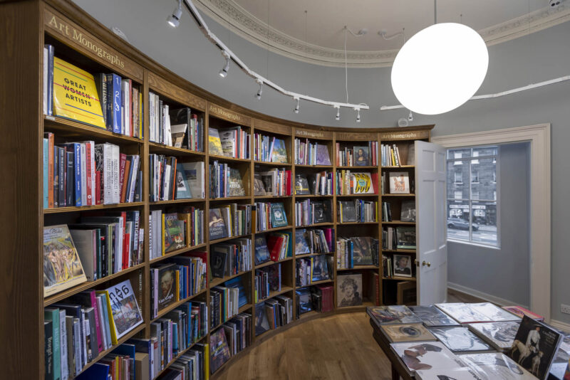 A view of a curved bookcase at Topping and Company, Edinburgh. Shelves filled with Art monographs. At the end of the shelf a curved door with a window behind, looking out onto the street.